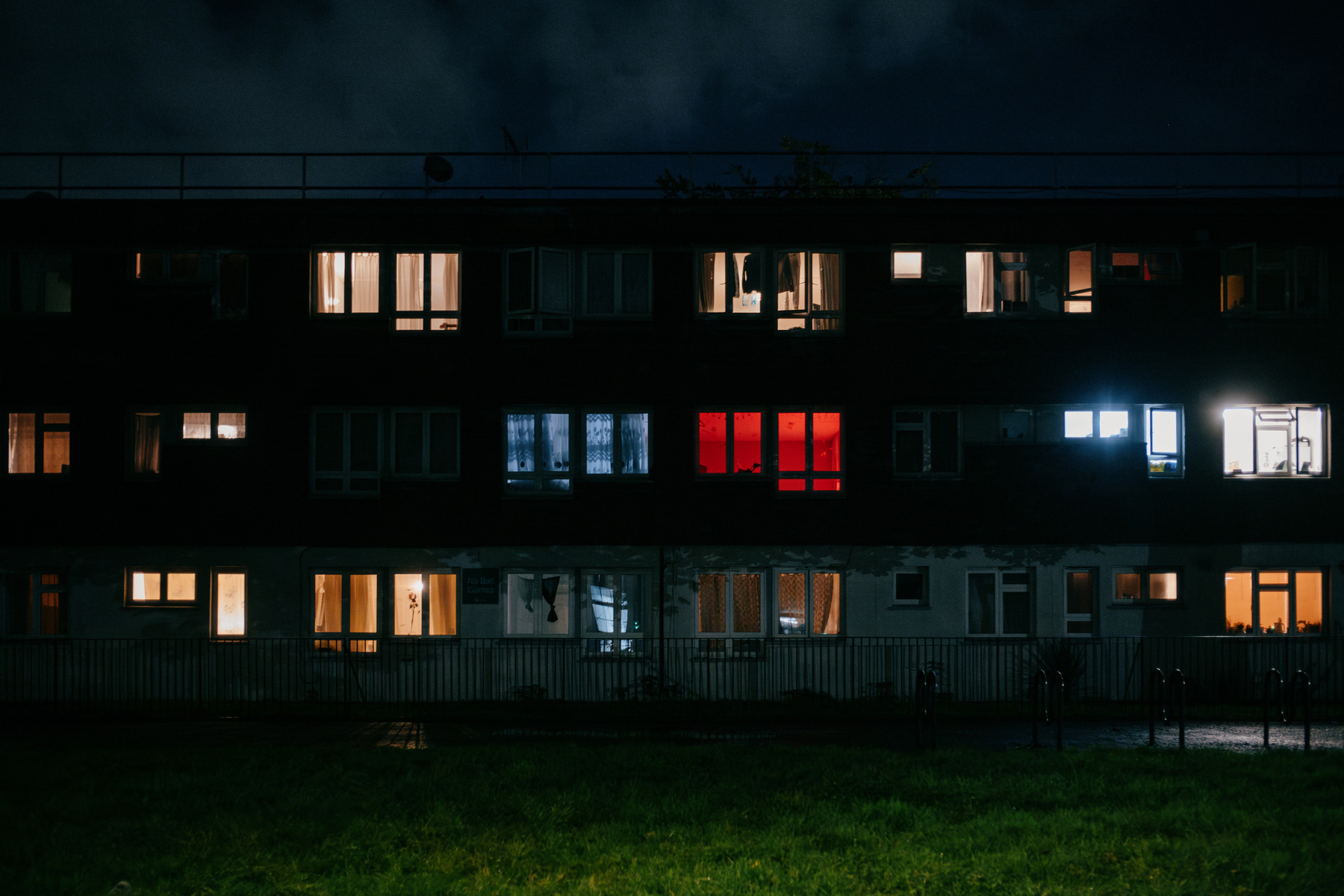 Side view of a low council block in north west London at night. The two central windows on the first floor are lit up, one red, one blue. The other windows are glowing with various warm to cool tones. The light from the windows is spilling over the patch of grass in front of the building.