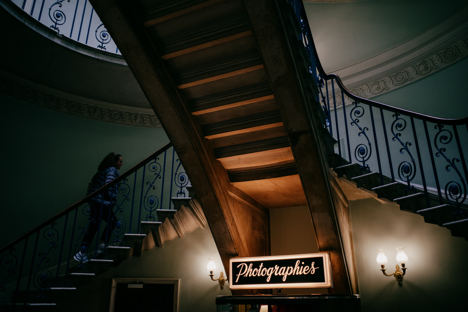 looking up at a narrow staircase running over the center of the frame. Curved staircases lead up to it from both sides with intricate ironwork railings. A woman is walking up the lefthand staircase. The top of a photo booth is seen above the bottom edge of the frame. It has a black illuminated sign with a white border and white text that reads 'Photographies'