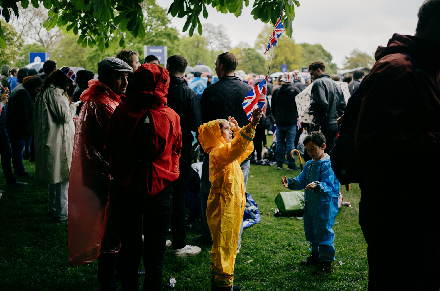 small child in a yellow rainsuit waving two small Union Jack flags at the Coronation of Kings Charles celebration in Hyde Park. The child is standing on grass, under the shelter of a sycamore tree, whose branches are just visible at the top of frame. There is a younger child in a blue rainsuit to the right, and two adults in red rain gear on the left. There is a large crowd surrounding the main subjects that extends into the distance.