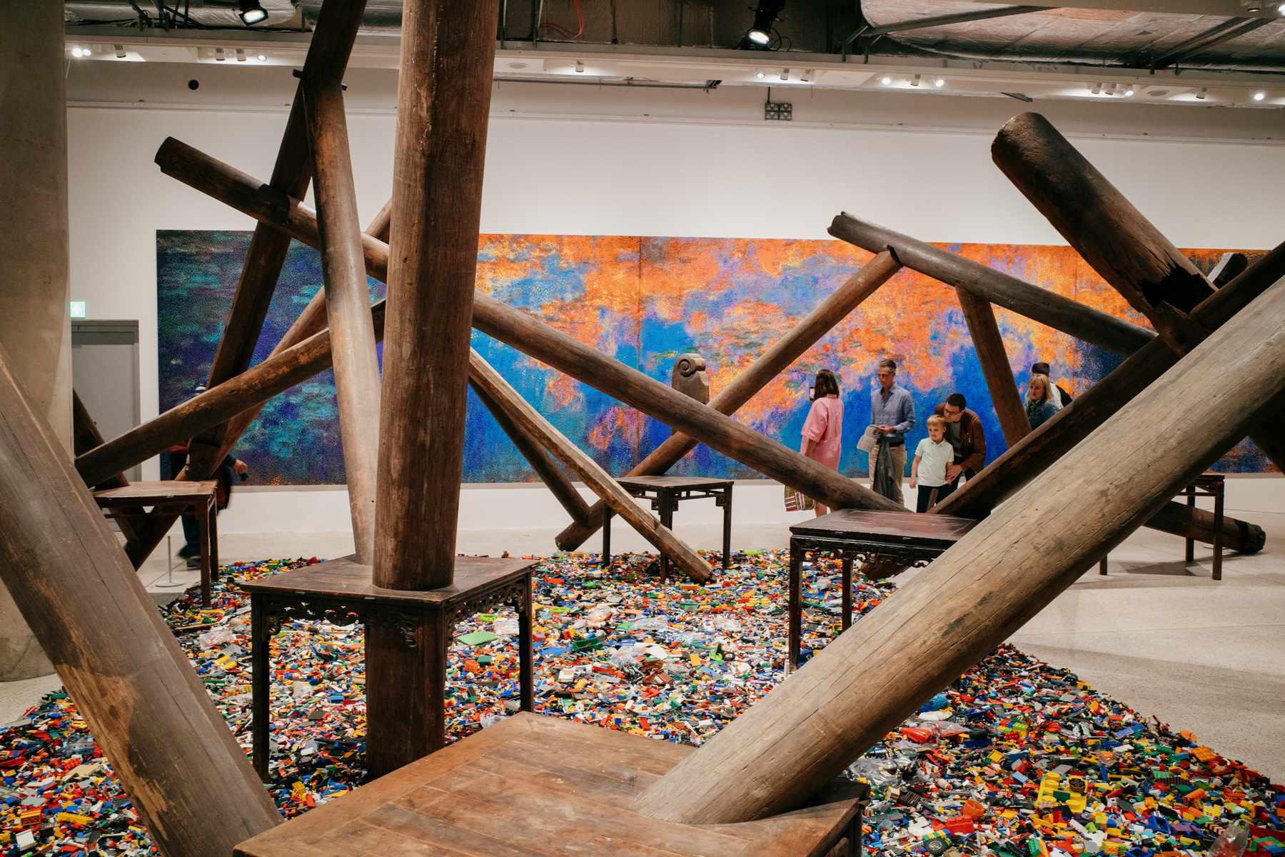 View through a large wooden sculpture by Ai Wei Wei in side a gallery. The wooden structure is made out of old furniture and beams from a temple. The foreground is covered with assorted multi-colored Lego pieces. On the far wall behind the sculpture there is a panoramic reinterpretation of Monet's Lilies painting made out of single Lego squares. There are a group of gallery visitors framed by the sculpture, standing in front of the Lego painting.