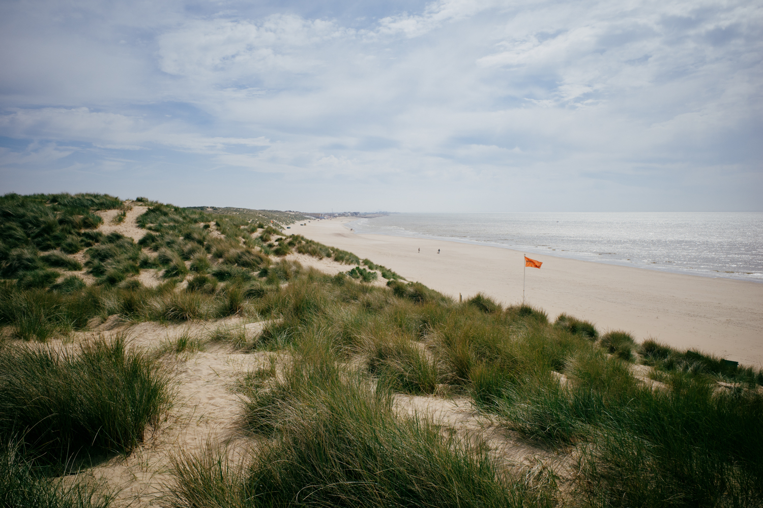 View down the beach at Camber Sands shot from the dunes. The grass and sand of the dunes are in the foreground and wrap around to the skyline on the left of frame. The strip of the sandy beach and the sea are to the right of frame. The sky is blue and there are some hazy clouds. There is an orange flag flying on a white flagpole in the distance in the center-right of the picture, at the edge of the dunes.