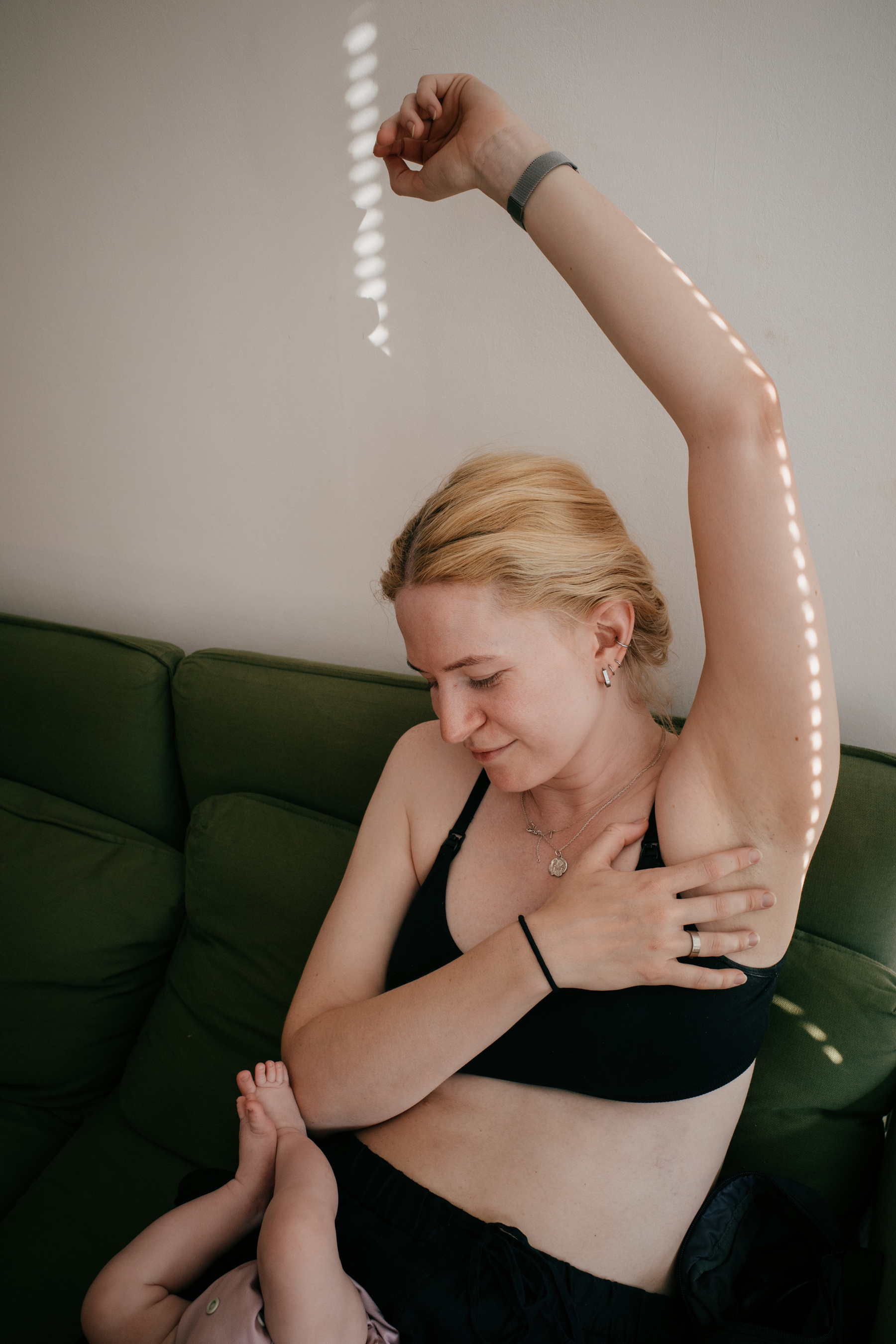 Woman sitting on a green sofa with her left arm held up. There is a line of spots of light running up her arm from gaps in the blinds. A baby's legs are sticking into frame from the bottom left corner. The woman is looking down at the baby