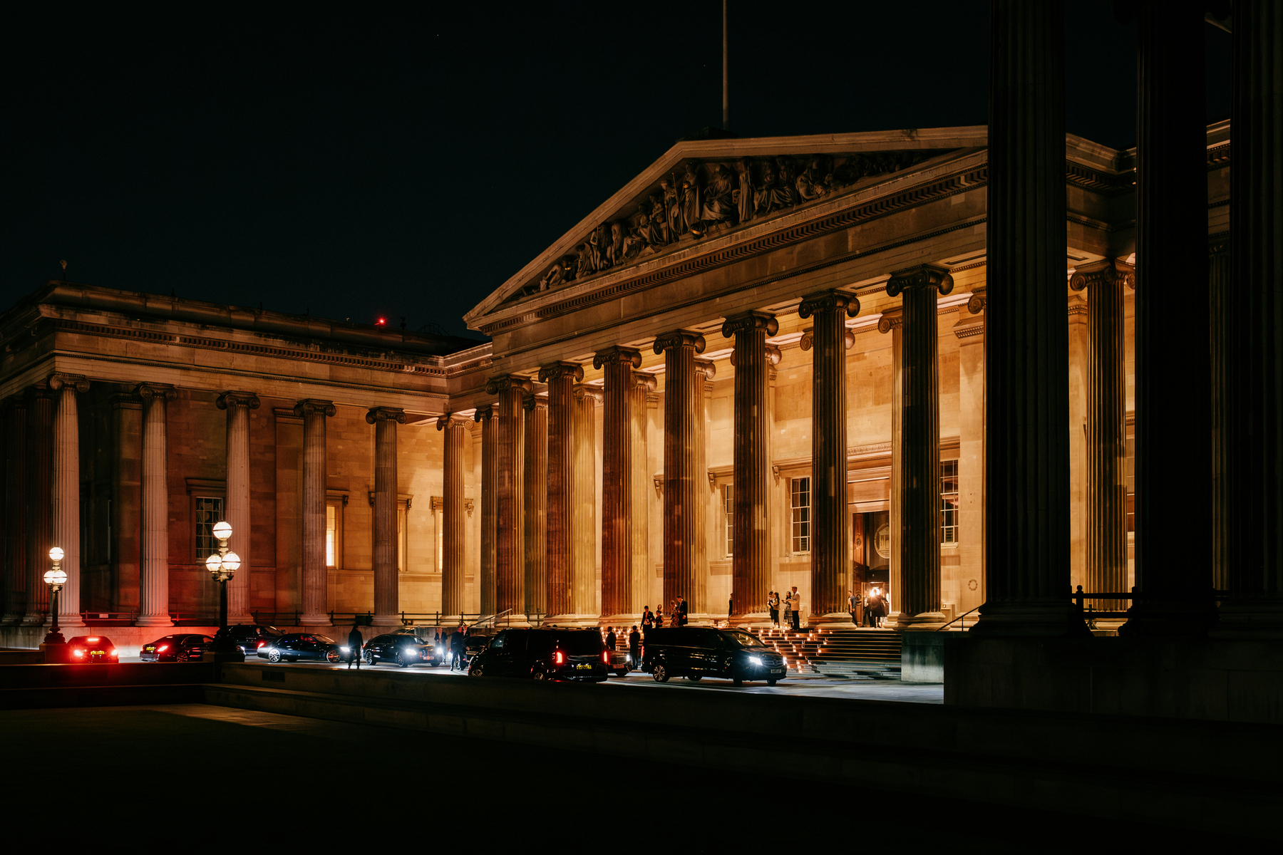 View of the British Museum main entrance at night. It is lit with warm light, there are candles on the steps and a line of cars waiting to pick up guests.