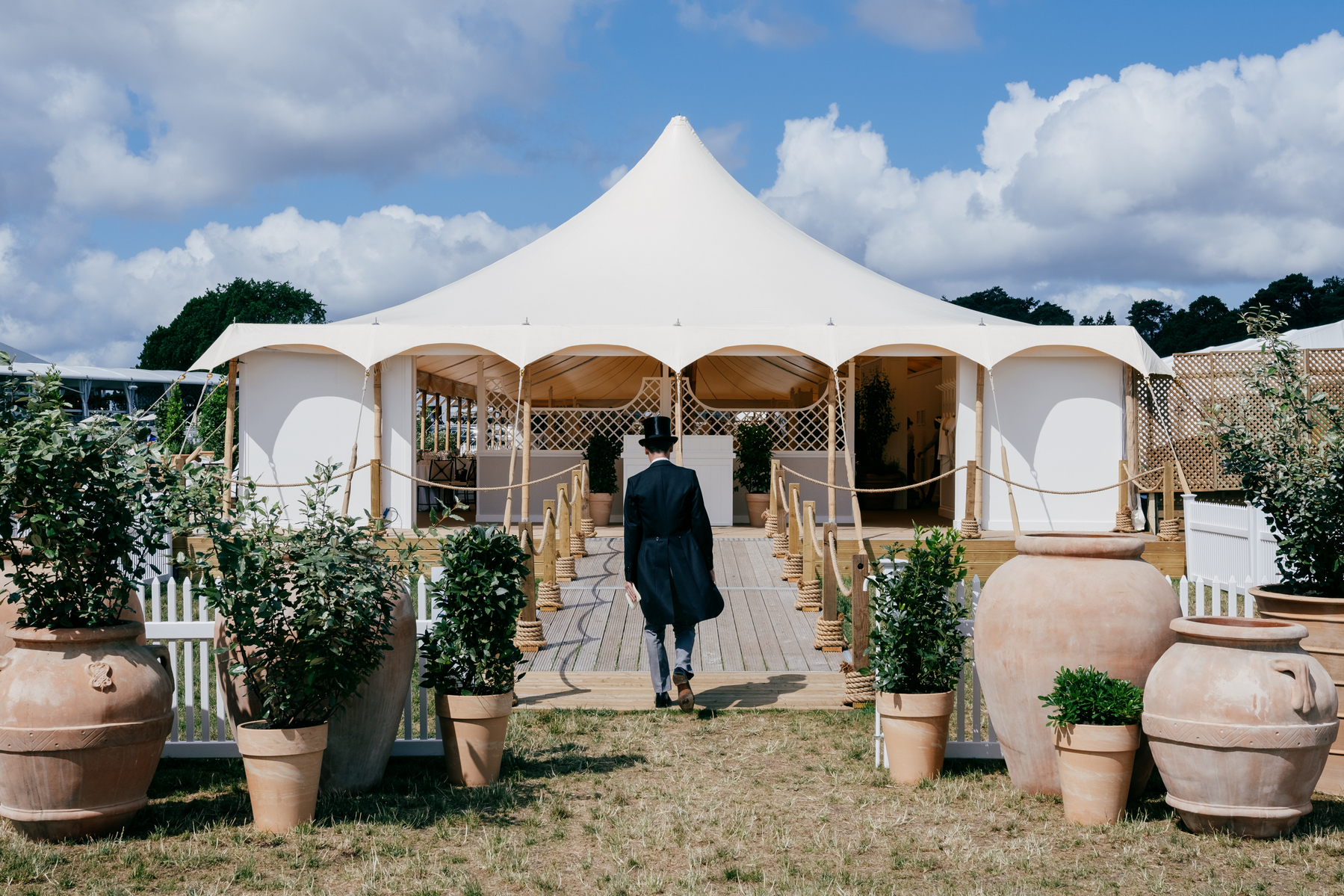 A man wearing top hat and tails walks up a shallow ramp into a marquee at Ascot. He is seen from behind and the ramp is flanked by large Roman style pots, some filled with ornamental shrubs. There are large cumulus clouds in the blue sky.