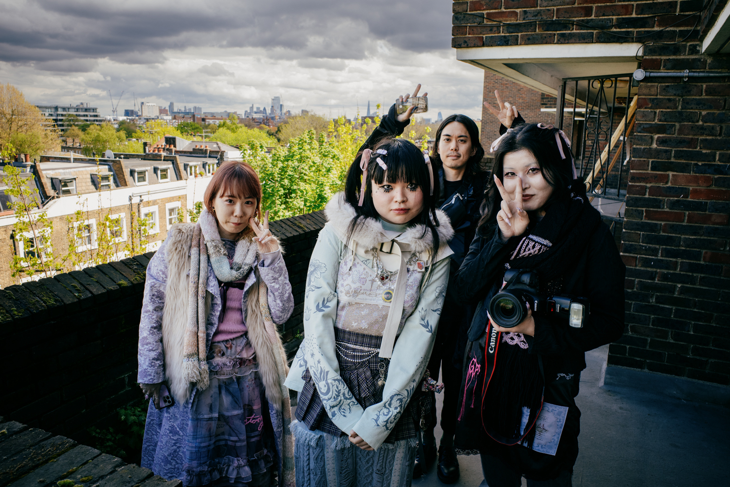 portrait of four Japanese fashion photoshoot crewmembers standing on an 5th floor open-air walkway in North London. Behind them, the view shows moody clouds and Canary Wharf on the skyline