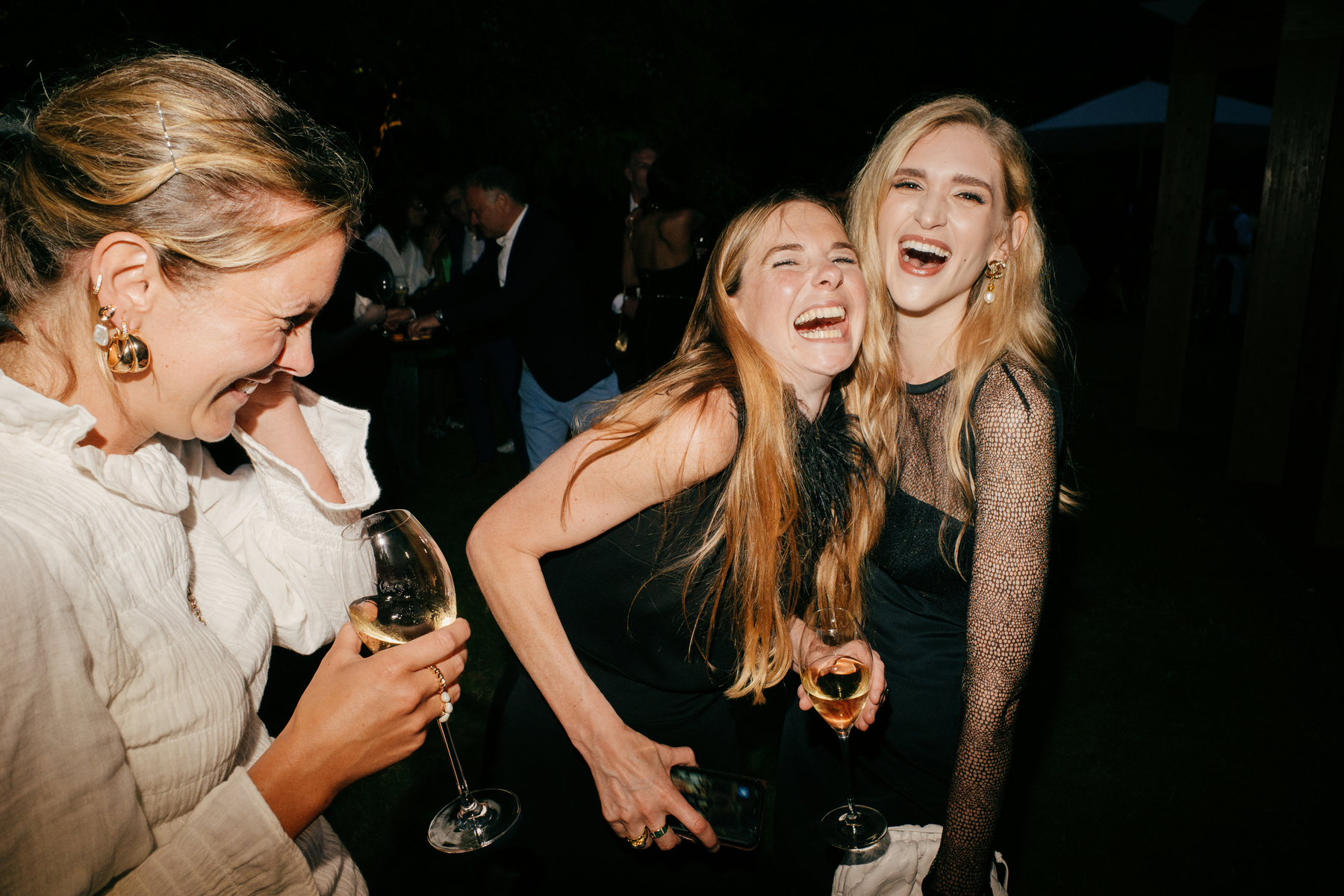 Flash on camera picture of two blonde women on right of frame laughing and hugging. There is another woman laughing at the left hand edge of frame