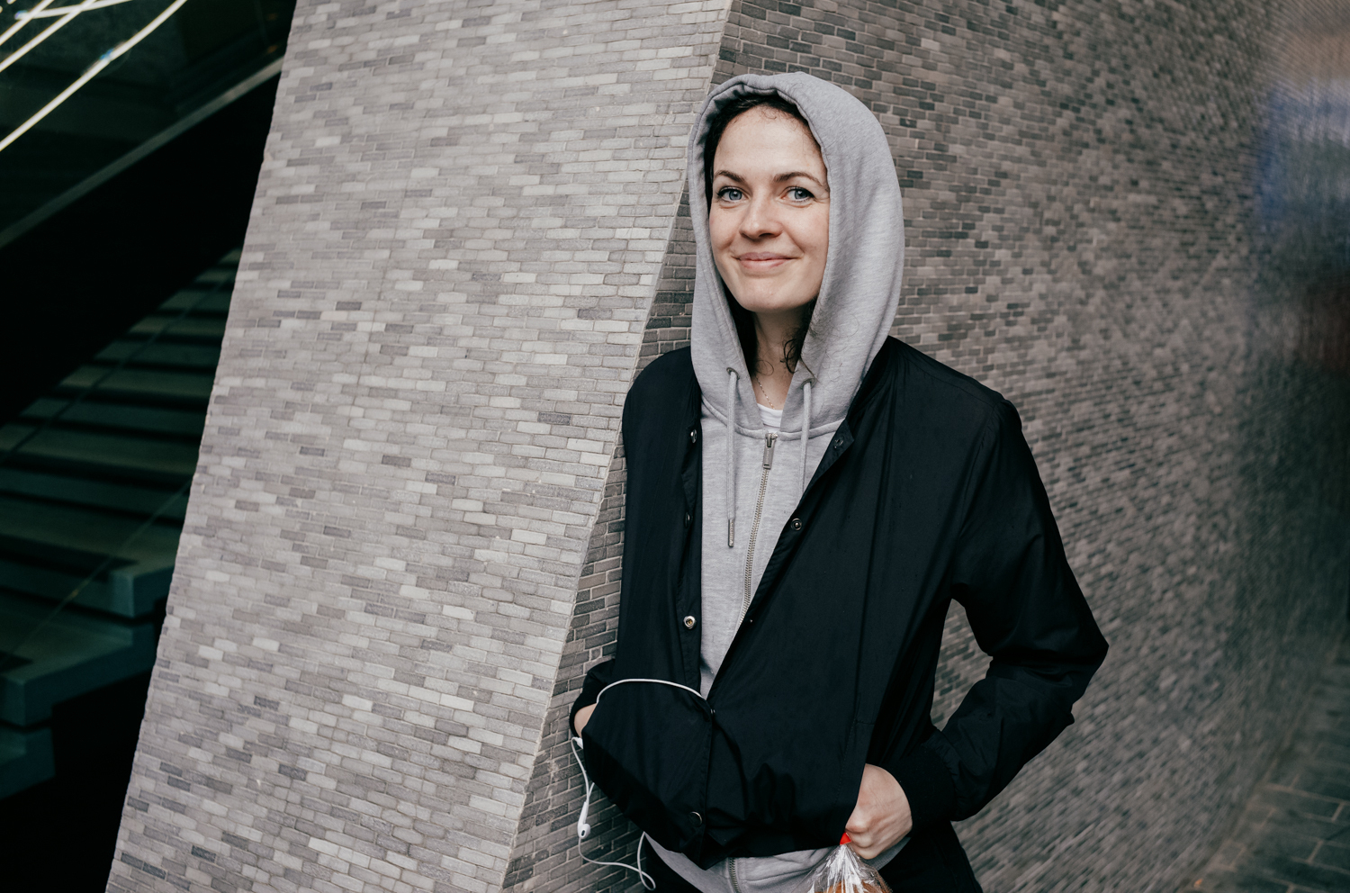 Portrait of actor, Georgia Winters, standing against a grey tiled wall against the base of Centre Point. She is wearing a black jacket and a light grey hoodie, with the hood up.