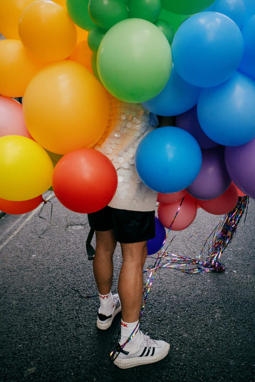 street photo of a man's legs with his upper body hidden by multi-colored balloons. The man is seen from the back and wearing white trainers and a white sequined top