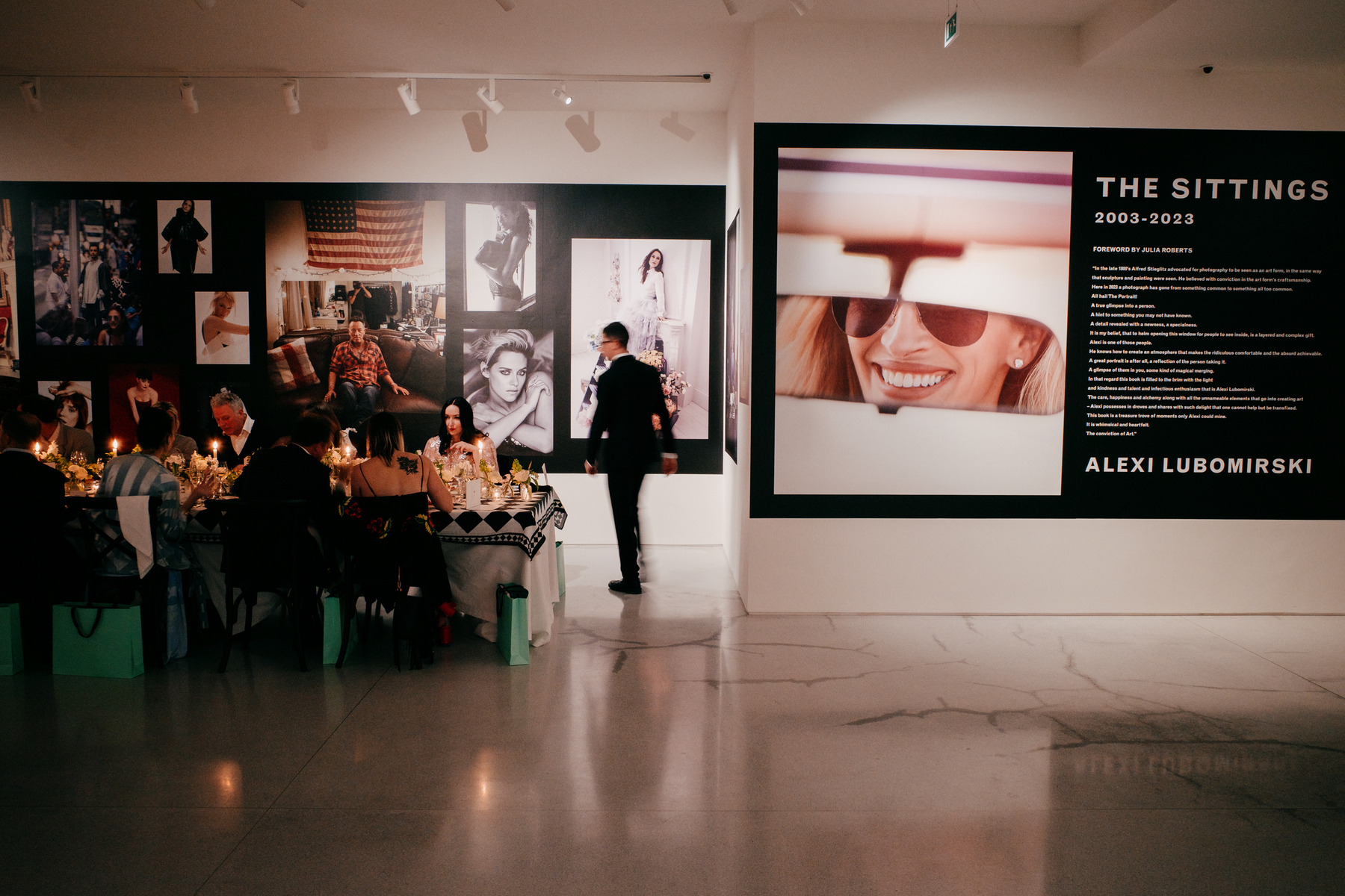 Private dinner in Claridges's Art Space for Alexi Lubomirski's book The Sittings. There is the end of a table entering the left of frame. It's lit by candles and guests are eating dinner. The space has a concrete floor and white walls. There are large photos printed mosiac style onto a black band that runs around the way of the room. There's a picture of Julia Roberts's smile reflected in a convertible's rearview mirror and the exhibition text on the wall to the right of frame.