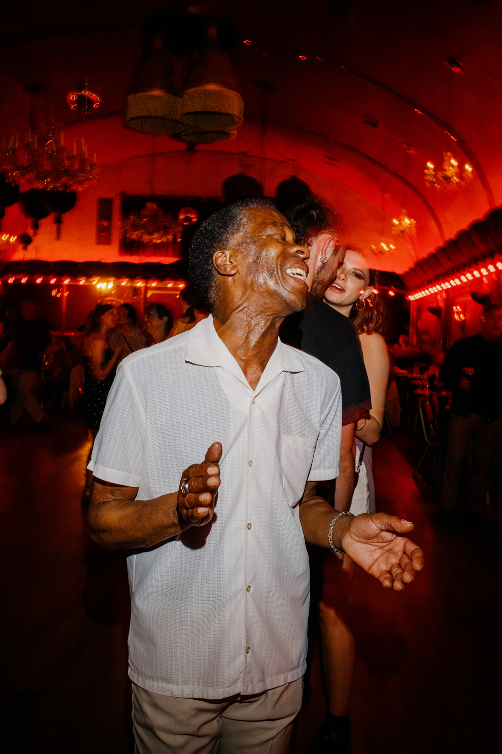 Man lit by flash dancing to northern soul at the rivoli ball room. He's wearing a short sleeved white shirt and his head is thrown back with his eyes closes. You can see a man and woman dancing in the background. She is looking to camera. The background is red from the ambient lighting. 