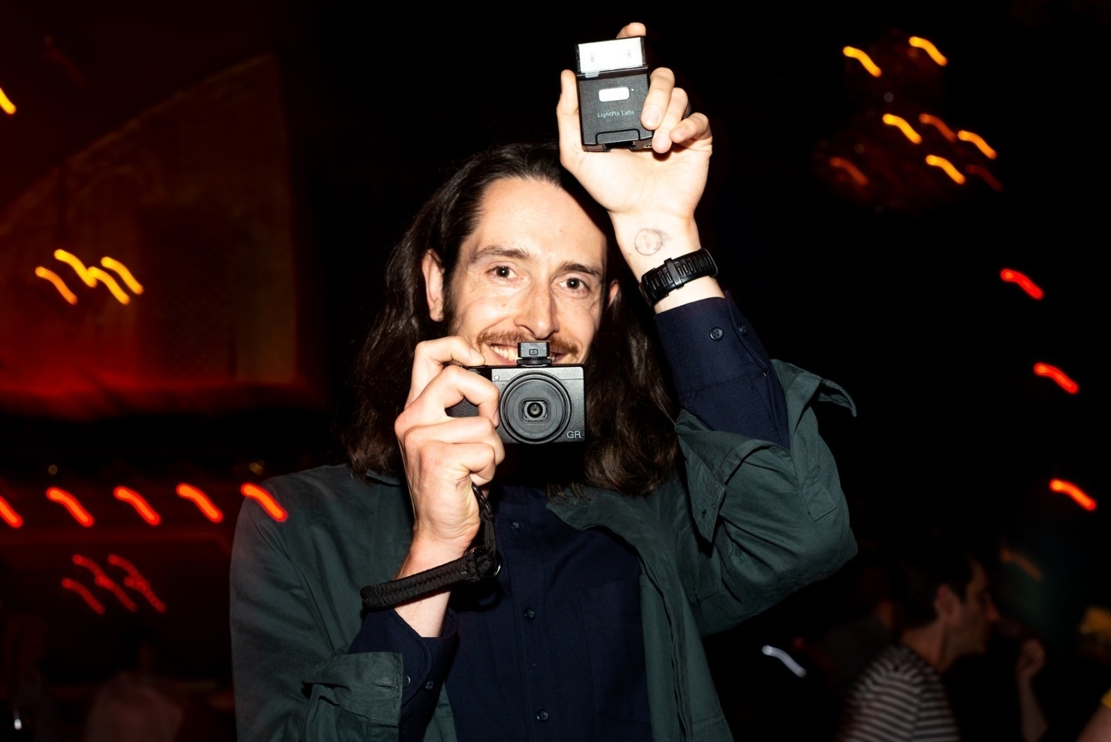 Oliver Holms holding up a Ricoh GRIII in his right hand and LightPix Labs FlashQ Q20II in his left hand. He has long brown hair, is wearing a dark green jacket and is smiling to the camera