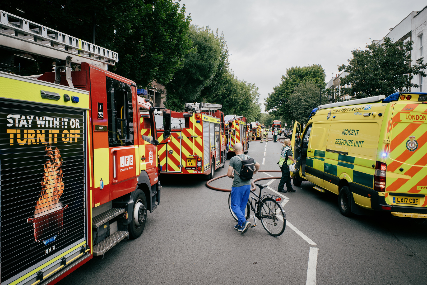 Four fire engines lined up the left hand side of Malden Road after an apartment fire. There is a man in blue trousers and green T-shirt walking his bike across the road, an ambulance on the right hand side, and various emergency personnel standing in the road surrounded by water hoses. There is a double decker bus waiting in the distance for the road to clear.