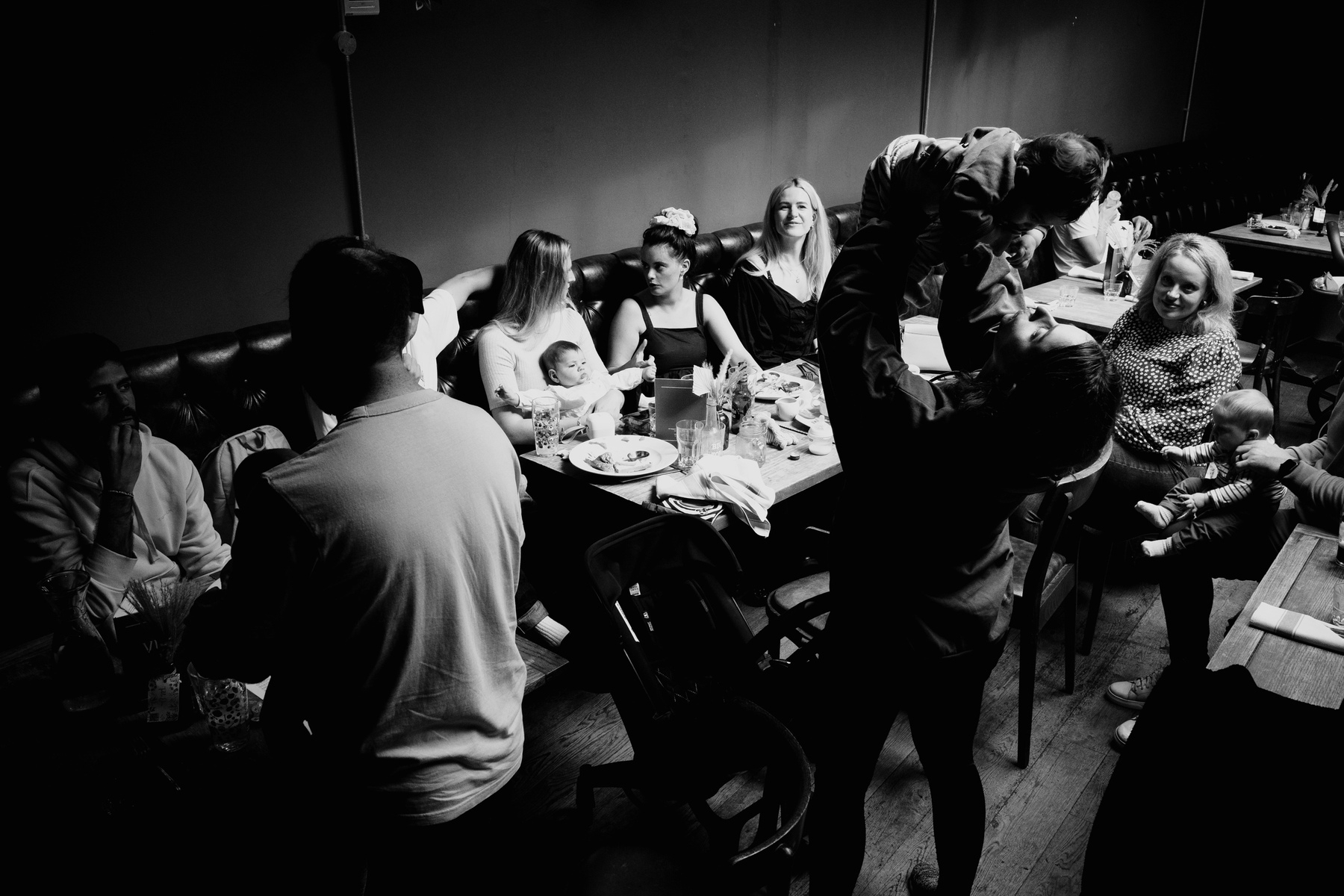 black and white photo of adults and babies seated around a long table in a pub. Most of the frame is dark and only the three women sat at the far side of the table are illuminated. In the foreground a silhouetted woman is holding her baby over her head and looking up at him