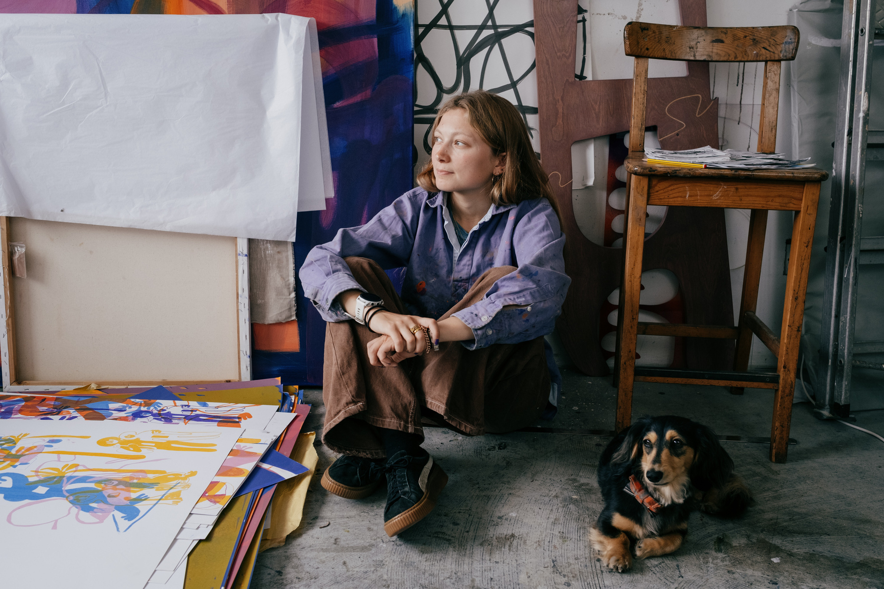 portrait of artist Alice Irwin in her studio. She sat on the floor and is looking to the left of frame. There are prints in a pile on the floor to her right, as well as paintings leaning against the wall. Her long haired dachshund is laying to her left, looking at the camera