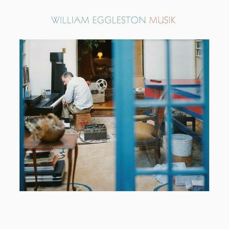 Cover of William Eggleston's Musik LP. The central photograph shows Eggleston sat at a low electric piano, with his back to the camera. There is a blue window pan on the right hand side of the frame that divides the picture in half