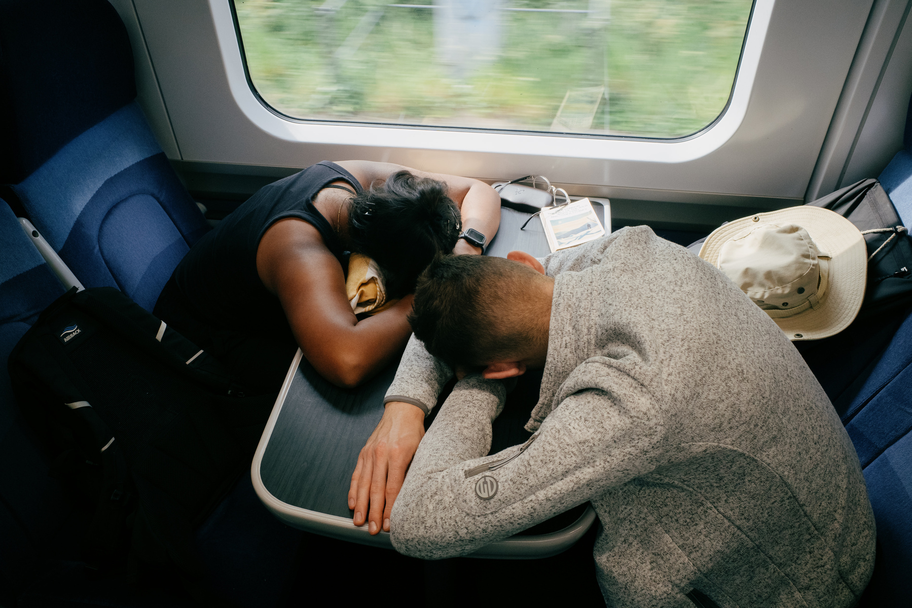 A man and a woman sleeping on a train with their heads in their arms on the table between them