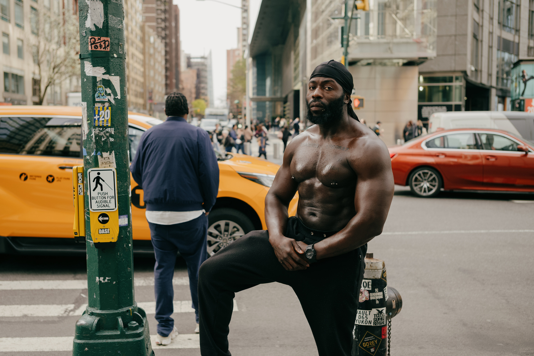 portrait of extremely muscular topless man with a beard and wearing a black doo-rag, posing next to a lamppost and fir hydrant. There is a road with an NYC yellow cab and a man crossing the street behind him on the left of frame.