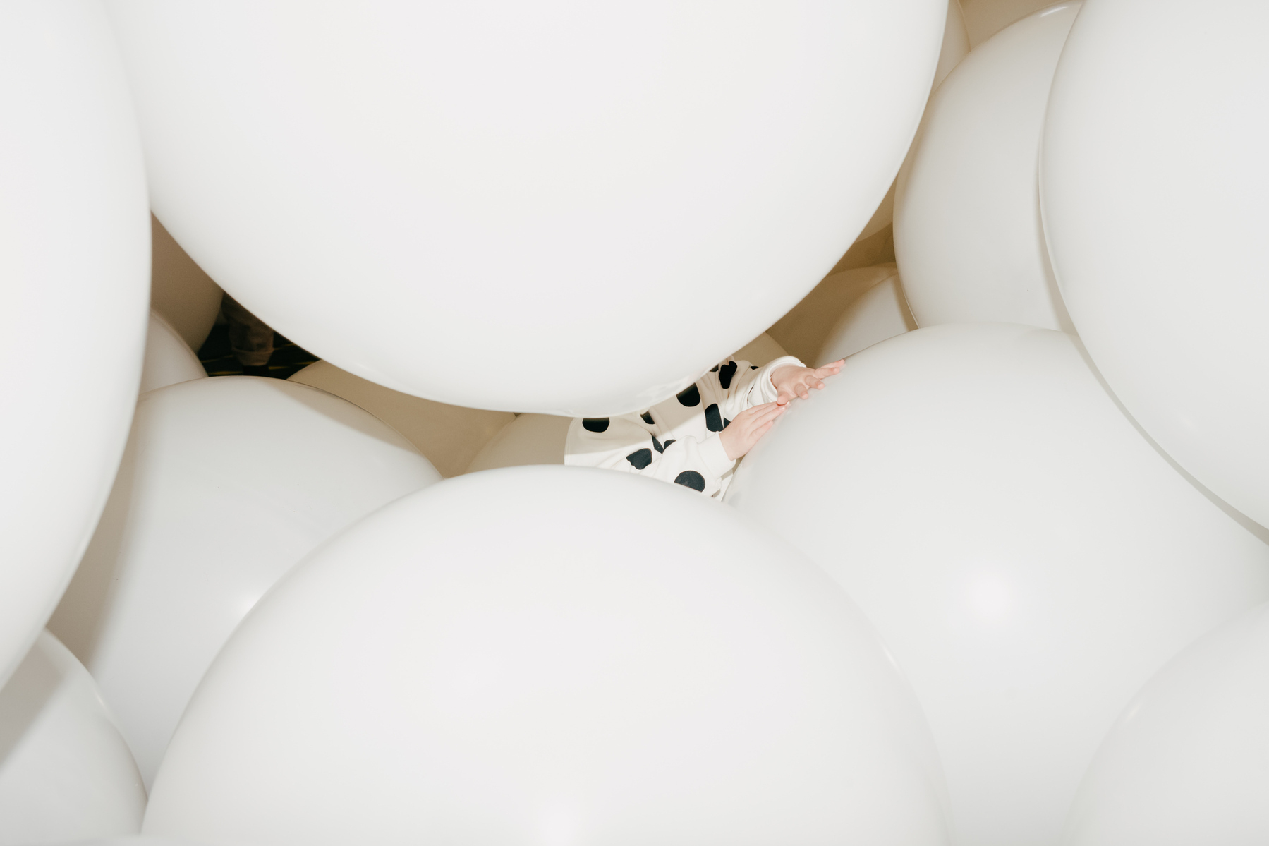 Child's hand seen in the gap between oversized white balloons. The child's jumper is white with black spots