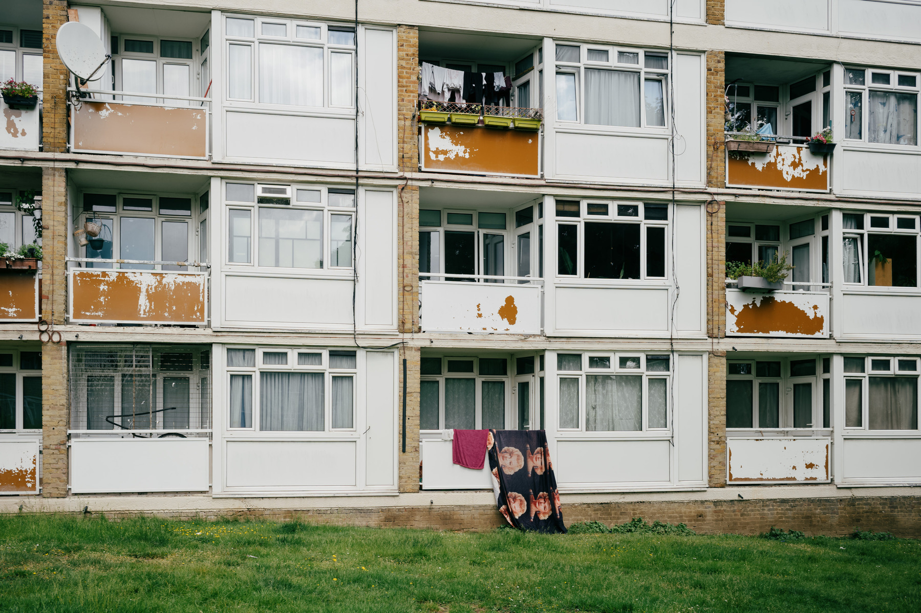 This image shows a weathered three-story residential apartment building with white and beige concrete panels, brown brick accents, and significant peeling paint revealing orange-brown patches underneath. The building features various window configurations, glass-enclosed balconies with personal items like plants and hanging laundry, and a patch of grass in the foreground, with an overall appearance suggesting mid-to-late 20th century social housing in need of maintenance. There is a black towel with a pattern of repeating Marilyn Monroe heads on a black background hanging over one of the balconies. 