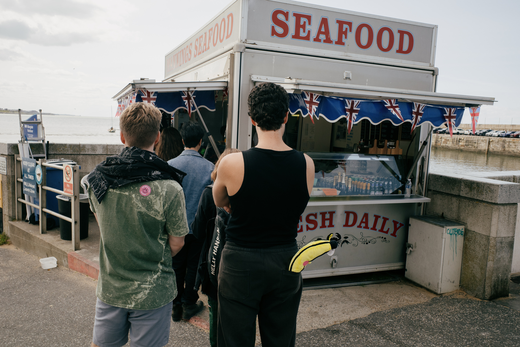 Two young men waiting at a seafood cart by Margate harbour. Both men are facing away from the camera. The man on the right is wearing black trousers and a black vest and has a large yellow banana soft toy sticking out of his right back pocket. 