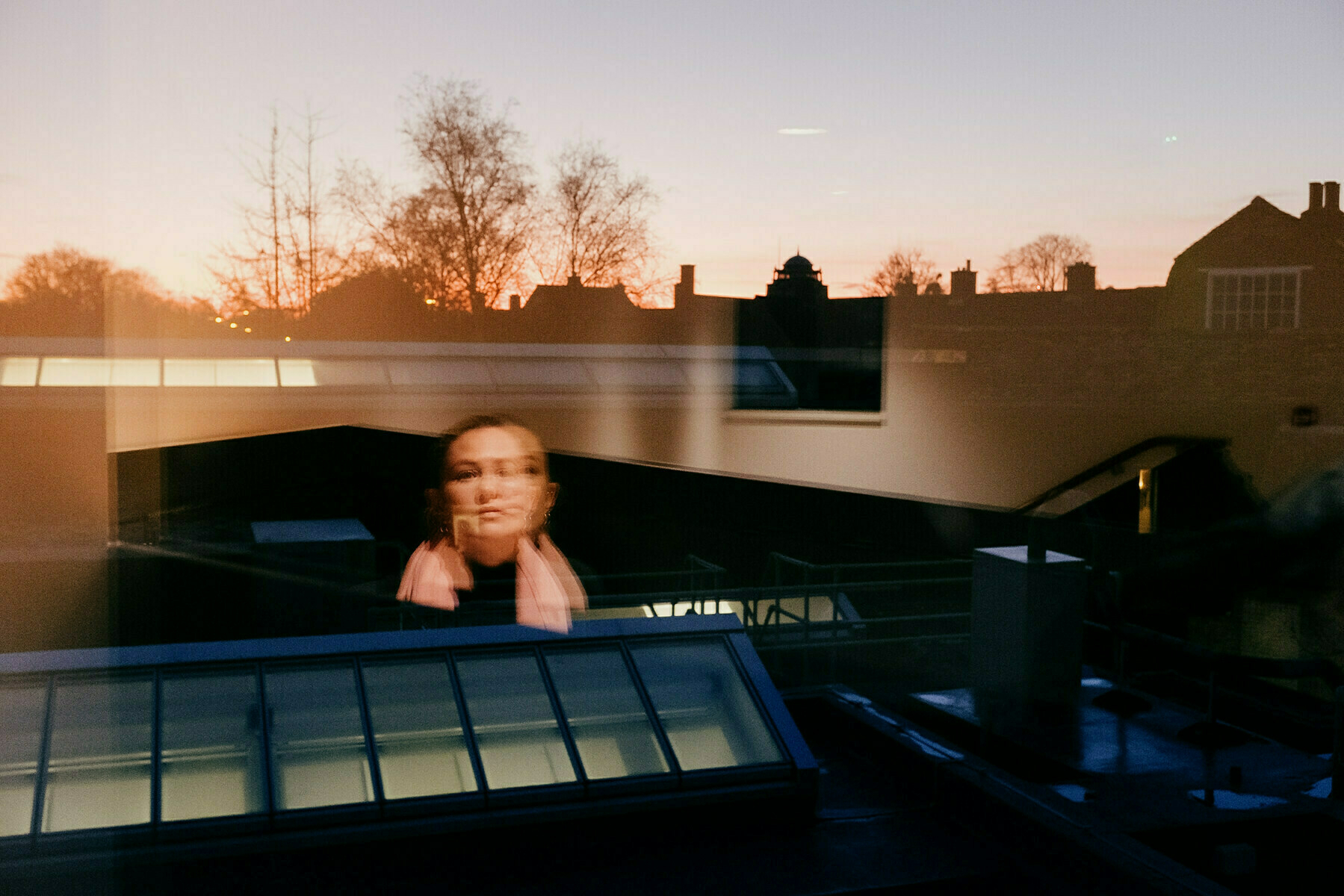 View out a window at dusk, looking over roof tops towards the treelined horizon. There is the reflection of a woman wearing a pink scarf floating in the left centre of frame. 
